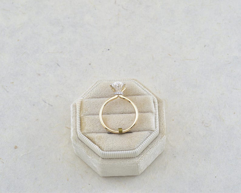 Puede incluir: Un anillo de oro con una piedra preciosa transparente de talla brillante redonda en un engaste de cuatro garras. El aro es fino y de oro pulido. El anillo est&aacute; colocado en posici&oacute;n vertical en un joyero octogonal de terciopelo beige.