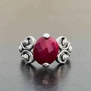 May include: A silver ring featuring a faceted, oval-shaped, deep red gemstone. The ring has ornate, swirling silver detailing on either side of the gemstone, creating a classic and elegant design. The ring is displayed on a dark surface.