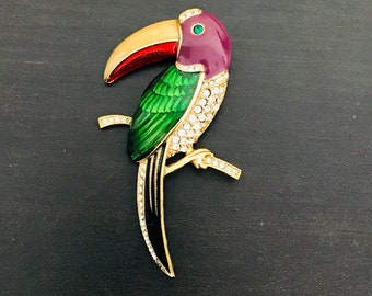 May include: A gold-tone toucan brooch with a red, green, and purple enamel finish. The toucan has a long, curved beak and is adorned with clear rhinestones.
