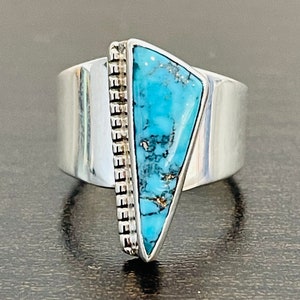May include: A silver ring with a turquoise stone set in a triangular shape. The stone is a vibrant blue with brown veins and is surrounded by a silver band with a decorative pattern.