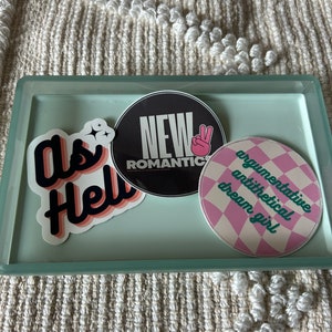 Könnte beinhalten: Ein hellblaues Tablett mit drei Aufklebern. Die Aufkleber sind rund und zeigen den Text "New Romantics", "Dream Girl" und "Argumentative Antithetical".