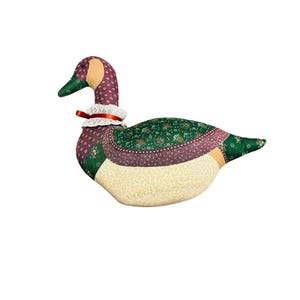 Puede incluir: Una figura de pato de tela rellena con un diseño de patchwork que presenta varios patrones florales y geométricos en tonos verde, morado y crema. El pato tiene un cuello de encaje blanco acentuado con una cinta roja delgada.