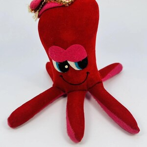 May include: A red plush octopus toy featuring a soft velvet-like texture, pink felt eyebrows, and large blue and white eyes. The octopus wears a small pink hat with gold trim on its head and has a stitched black smile on its face.