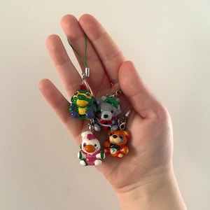 May include: Four miniature animal charms: a green frog, a gray bear, a white duck, and an orange bear. Each charm has a colorful design and is attached to a small string with a metal clasp. The charms are held in a hand.