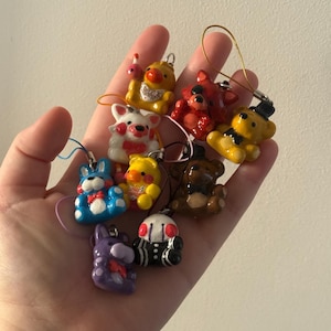 Puede incluir: Una colección de coloridos amuletos de personajes en miniatura. Los amuletos incluyen un conejo azul, un pato amarillo, un zorro rojo y un oso marrón, cada uno con detalles únicos. Están unidos a pequeños bucles para colgar.