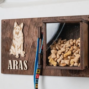 May include: Wooden dog treat dispenser with a clear window, a leash hook, and a cutout of a German Shepherd dog. The dispenser is labeled "ARAS".