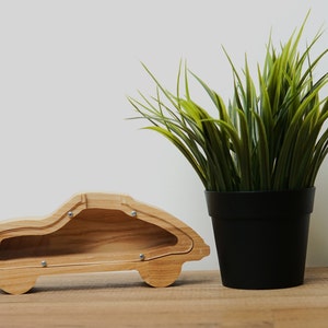 Può includere: Un salvadanaio di legno a forma di auto con uno slot nella parte superiore e un coperchio rimovibile. Il salvadanaio è su una superficie di legno accanto a una pianta verde in vaso.