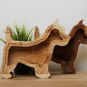 May include: Two wooden dog-shaped money boxes, one light brown and one dark brown, standing side-by-side on a wooden surface. A small potted plant is in front of the light brown money box.