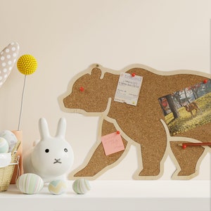 Peut inclure: Un tableau en liège en forme d'ours avec un billet, une photo et un post-it fixés dessus. Le tableau est sur une surface blanche avec un lapin blanc et des œufs de Pâques.