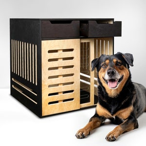 May include: A black and wood dog crate with a black dog sitting in front of it. The crate has two drawers and a wire door. The dog is looking at the camera and has its tongue sticking out.