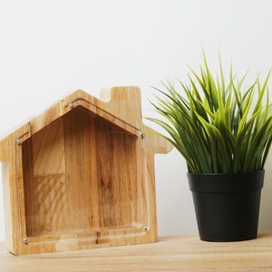 Può includere: Un salvadanaio di legno a forma di casa con un fronte in plastica trasparente. Il salvadanaio è appoggiato su una superficie di legno accanto a una pianta verde in vaso.