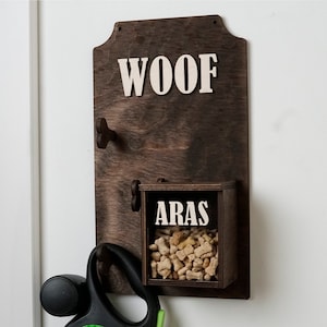 Pode incluir: Um dispensador de guloseimas para cães de madeira castanho escuro com a palavra "WOOF" no topo e a palavra "ARAS" na frente. O dispensador tem dois ganchos na lateral para as trelas e um gancho em forma de osso para uma coleira de cão. O dispensador está cheio de guloseimas para cães.