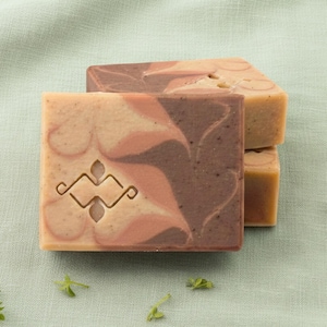 May include: Three square soaps with a swirled pattern in shades of brown, peach, and cream. The top soap has an etched decorative design. The soaps are stacked on a light green fabric, with sprigs of greenery scattered around.