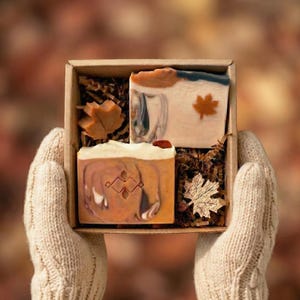 Peut inclure: Un coffret cadeau contenant deux savons faits à la main avec des motifs sur le thème de l'automne. Un pain de savon a une feuille d'érable et l'autre un motif tourbillonnant. La boîte est tenue par une personne portant des gants en tricot crème.