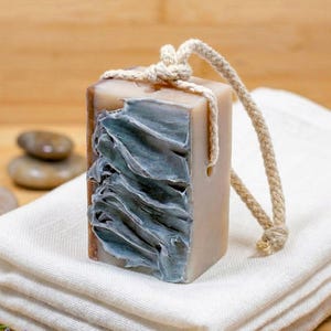 May include: A rectangular bar of soap with a textured, blue-grey design on one side. The soap is light brown with a rope attached for hanging. The soap rests on a white folded towel.