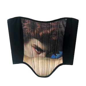 May include: A black corset featuring a print of a classical painting. The corset has a curved neckline and is constructed with vertical panels. The central panel displays a detailed image of a face with brown hair and an intense gaze.