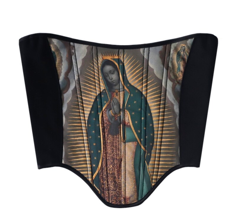 May include: A black corset with a colorful image of the Virgin Mary printed on the front. The image is surrounded by a gold halo and rays of light.