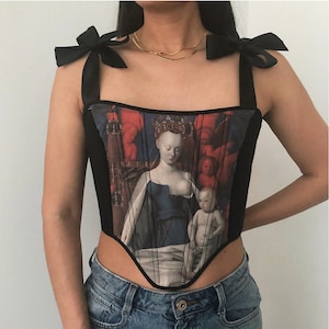 May include: A black corset with a print of a Renaissance painting depicting a woman breastfeeding a child. The corset has a pointed bottom and black ribbon straps that tie at the shoulders.