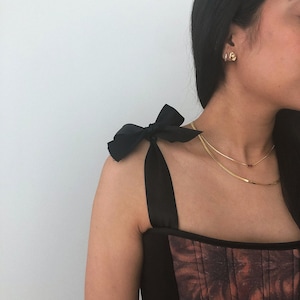 May include: Black top with shoulder ties and a patterned bodice. The top features a black bow on the shoulder and a gold necklace. The bodice has a brown and red floral pattern.