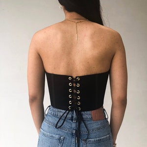 May include: A black strapless corset with gold-colored eyelets and a lace-up back. The corset is worn with blue denim jeans. The back of the corset is visible, highlighting the lacing detail.