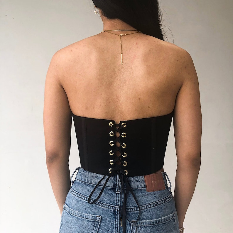 May include: A black strapless corset with gold-colored eyelets and a lace-up back. The corset is worn with blue denim jeans. The back of the corset is visible, highlighting the lacing detail.