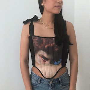 May include: Black corset top with a Renaissance painting print on the front. The top has thin black straps with bows on the shoulders. The corset features vertical lines and a curved bottom hem.