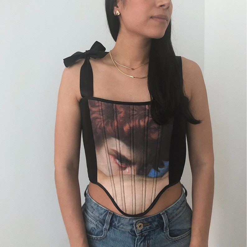 May include: Black corset top with a Renaissance painting print on the front. The top has thin black straps with bows on the shoulders. The corset features vertical lines and a curved bottom hem.