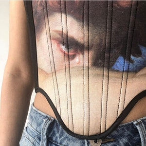 May include: A black corset top featuring a Renaissance painting print. The artwork depicts a close-up of a face with a tear. The corset has vertical black stitching and a curved neckline, paired with blue denim jeans.