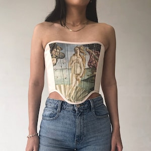 May include: A strapless corset with a pink background and a printed image of the Birth of Venus painting. The corset has boning and a pointed bottom.