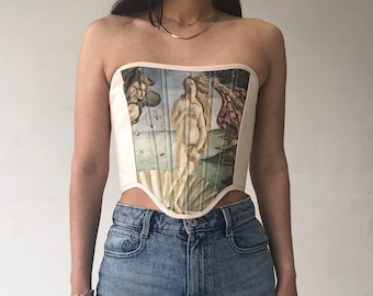 May include: A strapless corset with a pink background and a printed image of the Birth of Venus painting. The corset has boning and a pointed bottom.