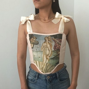 May include: A pink corset with a print of the Birth of Venus painting. The corset has white satin straps that tie into bows at the shoulders.