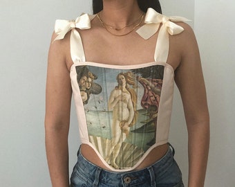 May include: A pink corset with a print of the Birth of Venus painting. The corset has white satin straps that tie into bows at the shoulders.