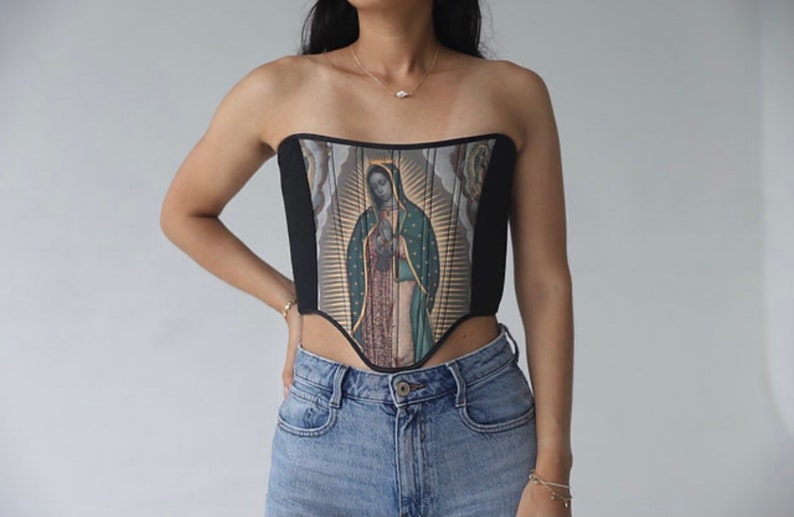 May include: A black and white corset with a colorful image of the Virgin Mary printed on the front. The corset has a pointed bottom and is worn over a pair of blue jeans.