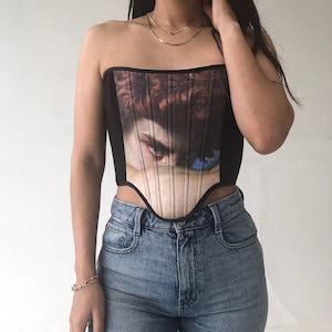 May include: Black strapless corset top featuring a Renaissance painting print of an eye. The corset has a structured bodice with vertical lines and is paired with blue denim jeans.