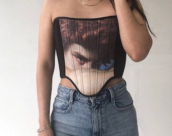 May include: A black corset with a print of a woman's face and eye. The corset has a boned structure and a pointed bottom.