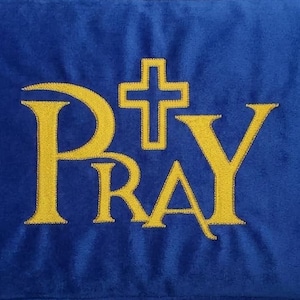 May include: A rectangular blue velvet pillow featuring the word PRAY embroidered in yellow thread. The letter A is stylized, and a yellow cross is positioned above the letter R, replacing the top portion of the letter to form the word PRAY.