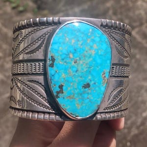May include: A wide silver cuff bracelet featuring a large, teardrop-shaped turquoise stone with dark matrix patterns. The silver band is decorated with intricate stamped leaf motifs, geometric lines, and notched edges along the top and bottom.