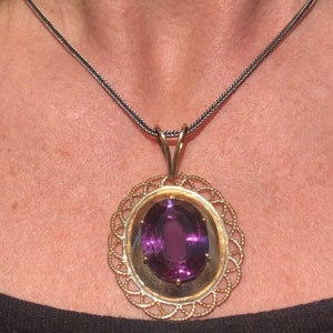 Puede incluir: Un collar con colgante de oro con una gran piedra preciosa púrpura de talla ovalada. La piedra preciosa está engastada en un marco de oro, rodeada por un delicado diseño de filigrana calada. El collar tiene una cadena oscura.