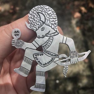 May include: A silver-toned metal brooch in the shape of a stylized figure. The figure has a horned head, holding a mirror in one hand and a bow and arrow in the other. The design features intricate etched details.