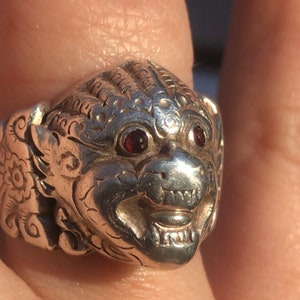 Op de afbeelding: Een zilveren ring met een gedetailleerde kop van een wezen. De ring heeft rode edelsteen ogen en ingewikkelde gravures. Het wezen heeft een grijnzende uitdrukking, met zichtbare tanden.