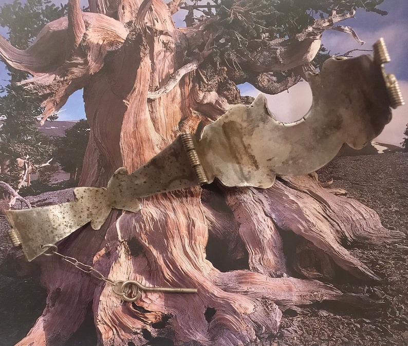 May include: A silver-toned metal belt with decorative cutouts and a key-shaped clasp. The belt features a chain and a hinged design. The metal has a worn, antique appearance. The background shows a tree trunk.