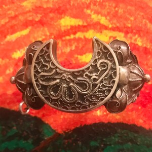 May include: A silver-toned filigree bracelet with a crescent-shaped centerpiece. The bracelet features intricate openwork designs and decorative spherical accents. The background is a colorful abstract painting with shades of orange, yellow, and green.