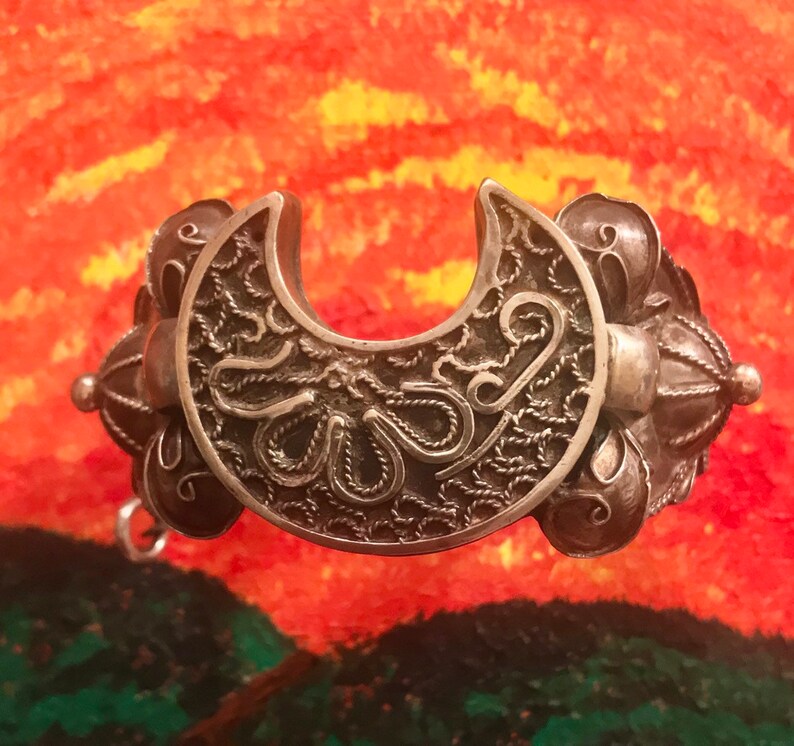 May include: A silver-toned filigree bracelet with a crescent-shaped centerpiece. The bracelet features intricate openwork designs and decorative spherical accents. The background is a colorful abstract painting with shades of orange, yellow, and green.