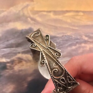 May include: A silver-toned filigree bracelet with an ornate cross design. The bracelet features intricate scrollwork and a small ring at the end. The background is a blurred image of a seascape.