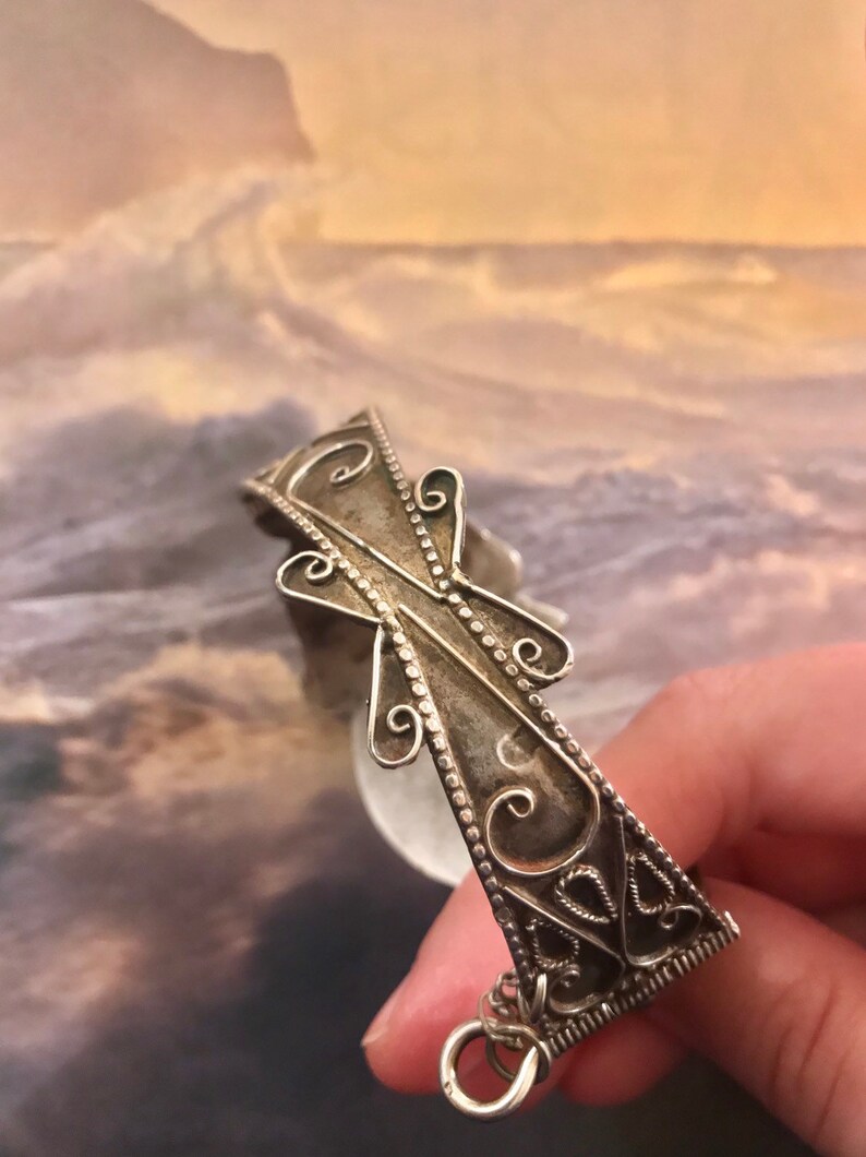 May include: A silver-toned filigree bracelet with an ornate cross design. The bracelet features intricate scrollwork and a small ring at the end. The background is a blurred image of a seascape.