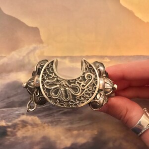 May include: Silver-toned bracelet with a crescent-shaped centerpiece. The bracelet features intricate filigree and floral designs. The bracelet is held by a hand, showcasing the detailed craftsmanship of the jewelry.