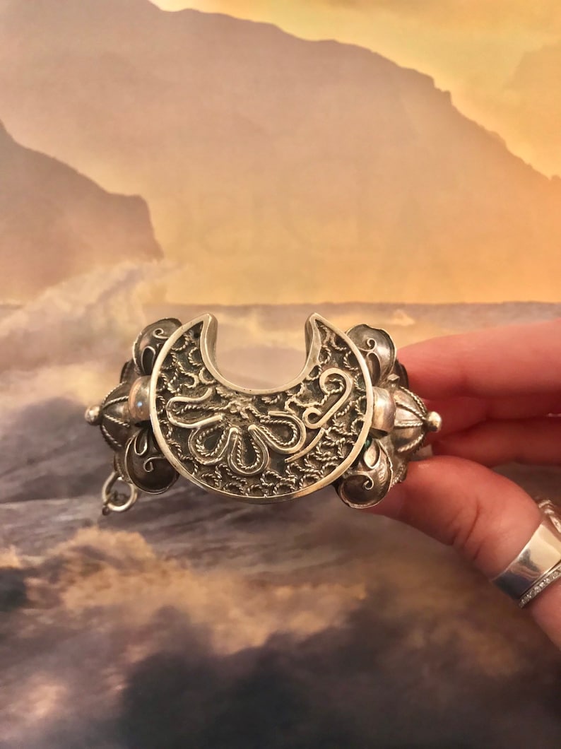 May include: Silver-toned bracelet with a crescent-shaped centerpiece. The bracelet features intricate filigree and floral designs. The bracelet is held by a hand, showcasing the detailed craftsmanship of the jewelry.