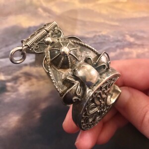 May include: An antique silver-toned ring with intricate filigree and decorative elements. The ring features a detailed design with a central raised element and a textured surface. The ring has a loop at the top and a decorative band.