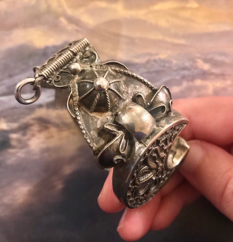 May include: An antique silver-toned ring with intricate filigree and decorative elements. The ring features a detailed design with a central raised element and a textured surface. The ring has a loop at the top and a decorative band.