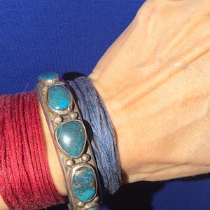 May include: A silver cuff bracelet featuring five polished, oval-shaped turquoise stones with dark matrix patterns. The bracelet is worn alongside a dark blue thread wrap and a red thread wrap, showcasing a metallic band with small silver bead accents.